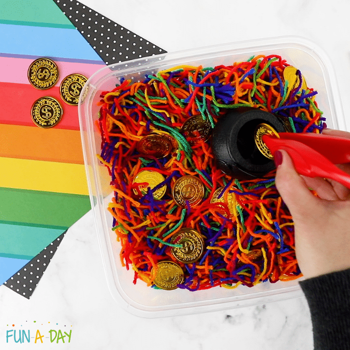 close up of a rainbow yarn sensory bin filled with gold coins and a hand putting a coin into a small black pot