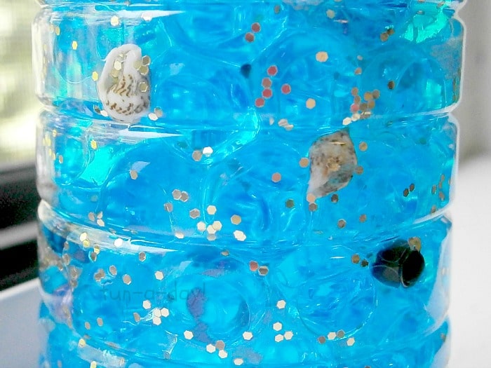closeup of blue water beads, tiny shells, and gold glitter in a pirate treasure bottle for kids