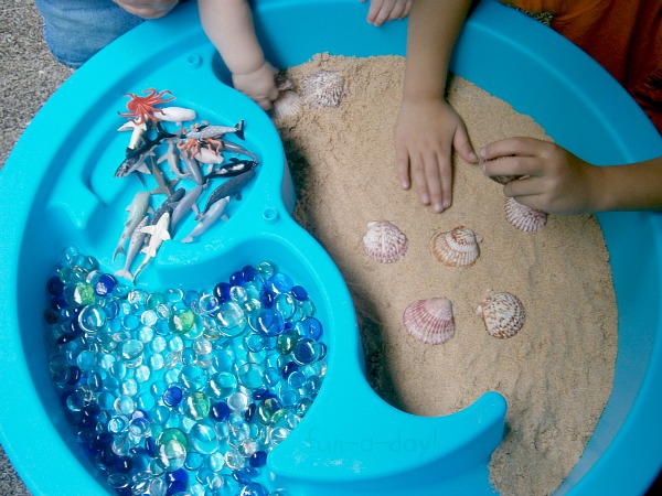 children's hands in a sensory table filled with play sand, glass gems, shells, and ocean animal toys