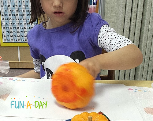 a girl rolling a pumpkin covered in paint onto white construction paper to create pumpkin process art