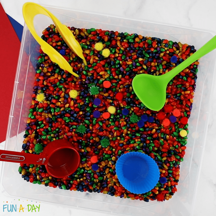 image of colored popcorn kernels in a sensory bin with tweezers and spoons