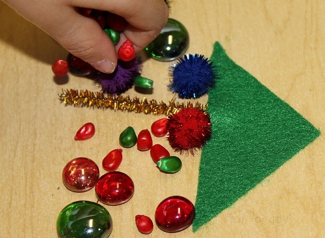 Child adding colored corn kernels to contact paper, along with green felt, glass beads, and sparkly pipe cleaners.