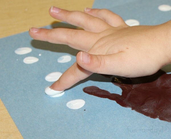 child using her fingers to paint white snowflakes around a winter hand print tree