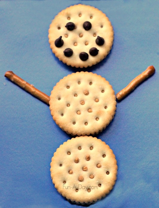 three peanut butter cracker circles on top of each other representing a snowman body with pretzel arms and a chocolate chip smile