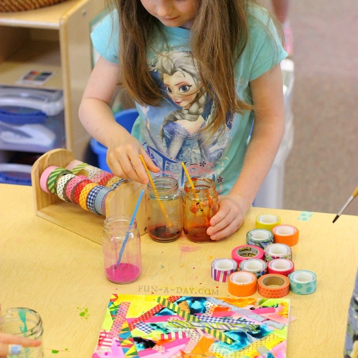 Preschool child creating tape and watercolor canvas art surrounded by liquid watercolors in jars and colorful washi tape.