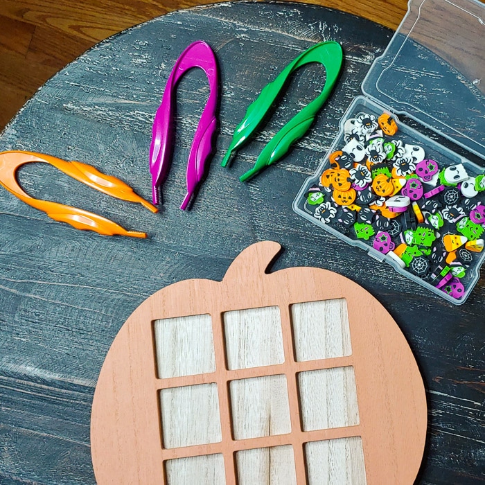 Pumpkin shaped tic-tac-toe board with three kid tweezers nearby and Halloween tiny erasers