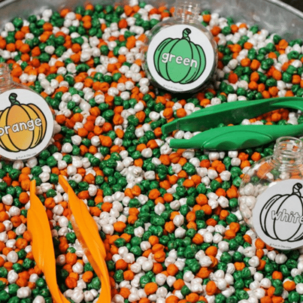 Tray of dyed chickpeas with kid tweezers and three plastic bottles labeled orange, green, and white.