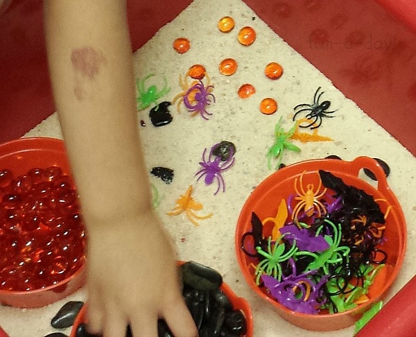preschooler playing in sand sensory bin that includes orange glass gems, spider rings, and bat rings
