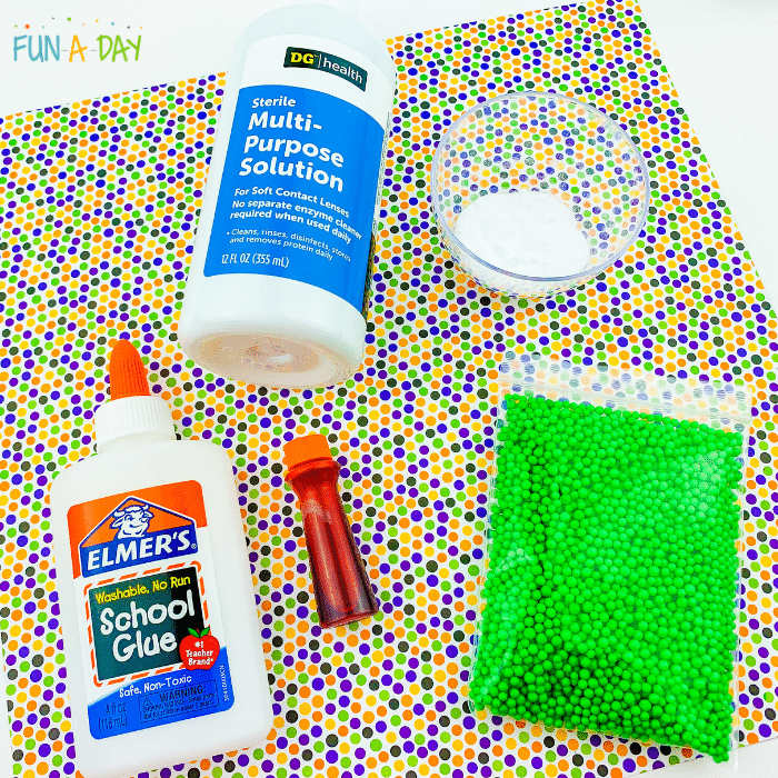 Ingredients for activity which includes contact lens solution, Elmer's school glue, orange food coloring, pack of green foam beads, and a ramekin of baking soda all laid out on a multicolored polkadot craft mat.