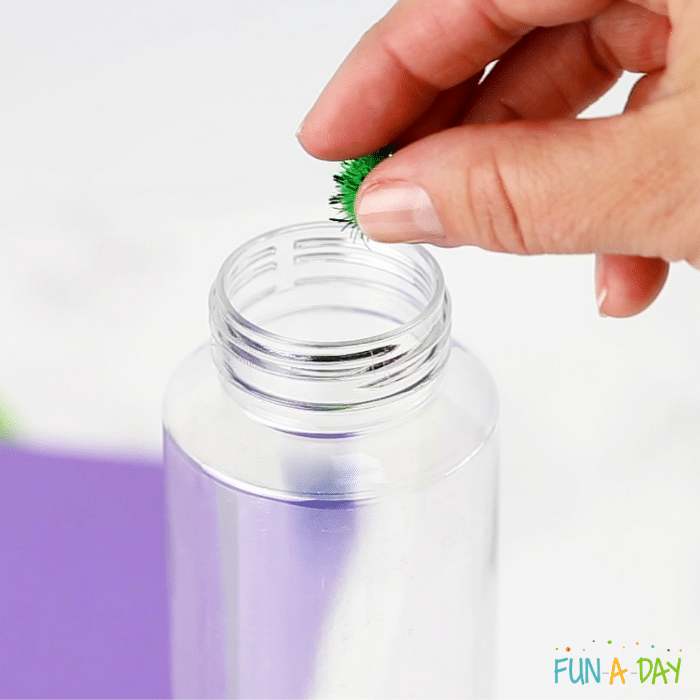 Hand placing green pompom into clear bottle.