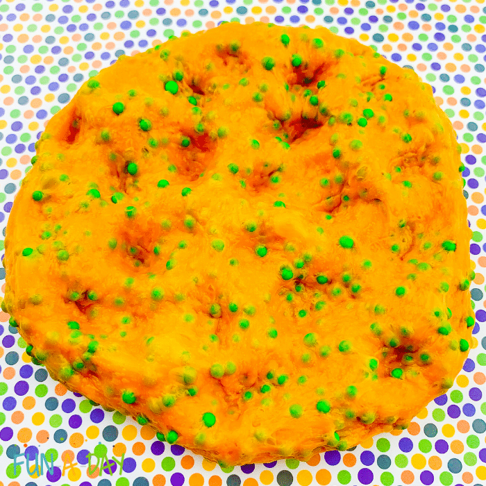 Orange slime with green foam beads with several finger indentions on a multicolored polka dot crafting mat.