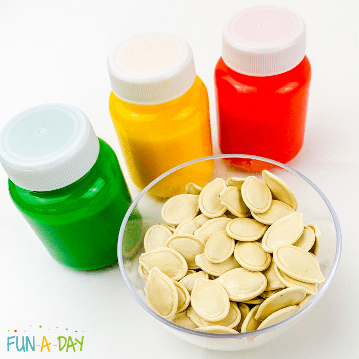 Green, yellow, and red bottles of paint and a small bowl of pumpkin seeds as ingredients for fall painting for kids.