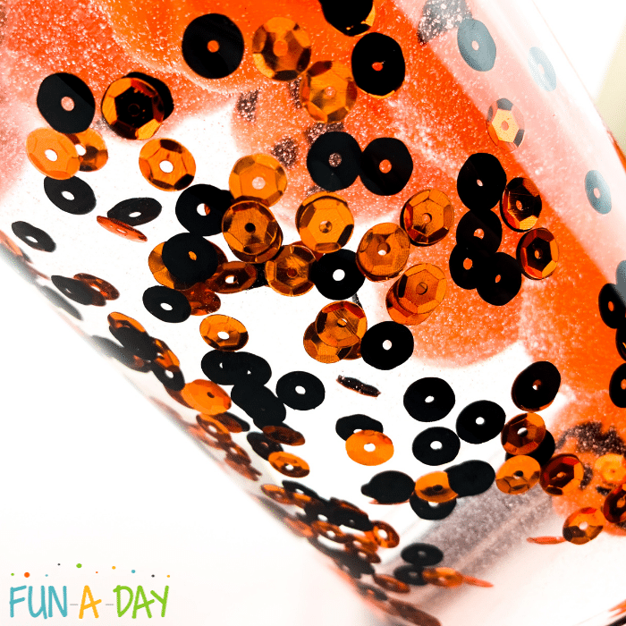 A clear bottle of range and black sequins and jack-o-lanterns floating in a clear bubbly liquid.