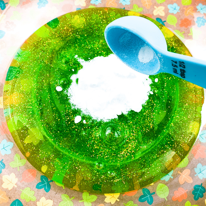 Half a tablespoon baking soda being poured into large portion of green glitter glue.
