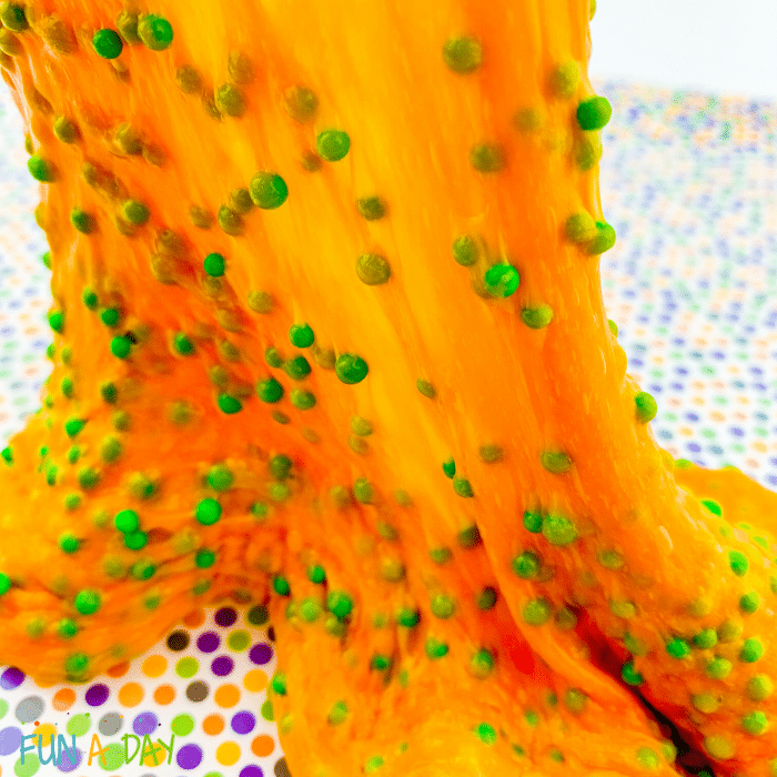 Orange slime with green foam beads being stretched out from a multicolored polkadot craft mat.