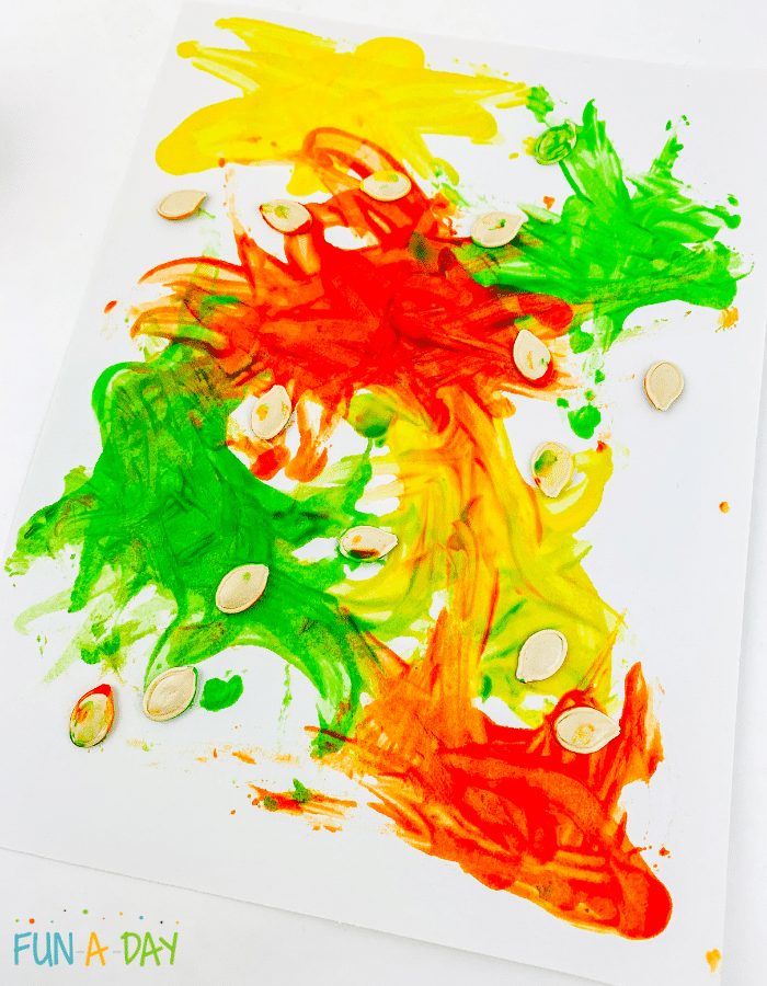 Red, yellow, and green paint being swirled with pumpkin seeds.