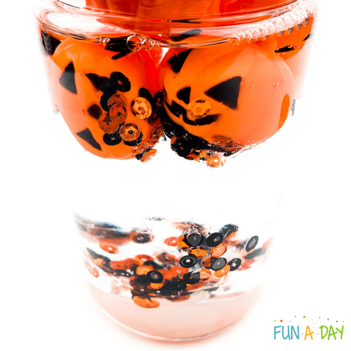 Clear bottle with orange and black sequins and small plastic jack-o-lanterns floating in a clear liquid.
