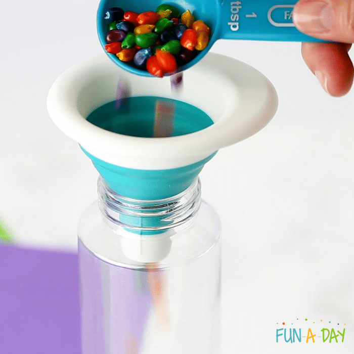 Tablespoon full of colorful corn kernels being poured into a clear bottle with a funnel.