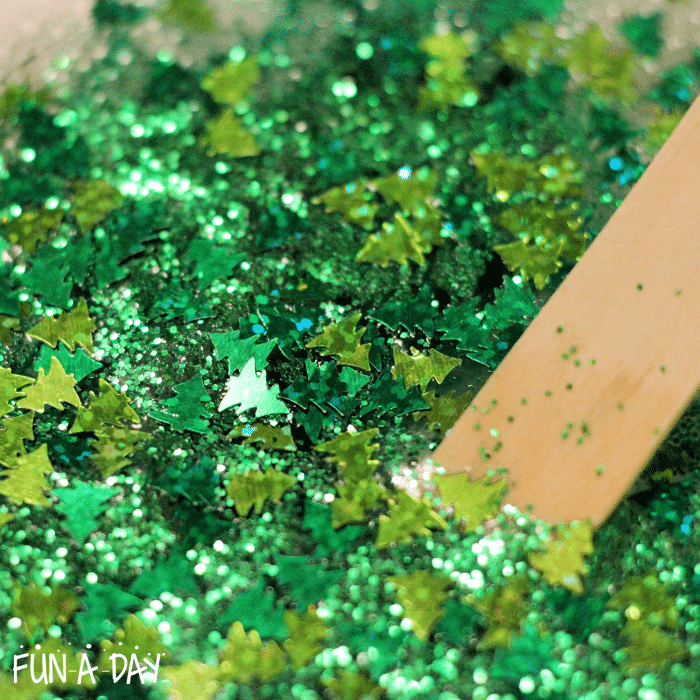 Tree confetti and green glitter being mixed with glue using a craft stick.