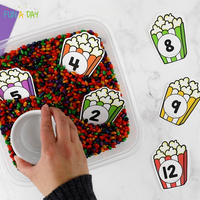 Bin of multicolored popcorn kernels with number cards both inside and lined up next to the bin. A small bowl being placed inside the container of corn.