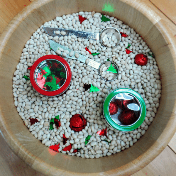 Christmas sensory bin with white navy beans with red jingle bells, Christmas tree confetti, measuring spoons, and cups scattered throughout the beans.