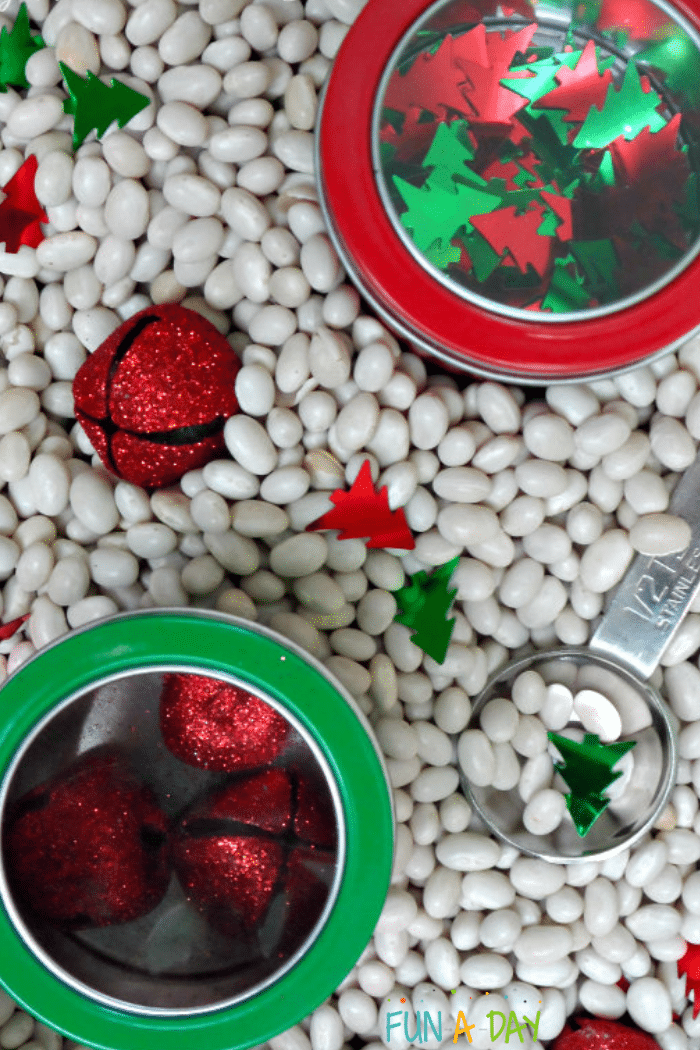 Christmas sensory bin with red jingle bells, Christmas tree confetti, and measuring spoon scattered throughout white navy beans.