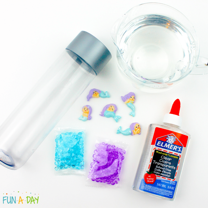 Materials needed for the mermaid sensory bottle which include clear sensory bottle, mermaid charms, blue fish bowl beads, purple fish bowl beads, Elmer's clear glue, and water.