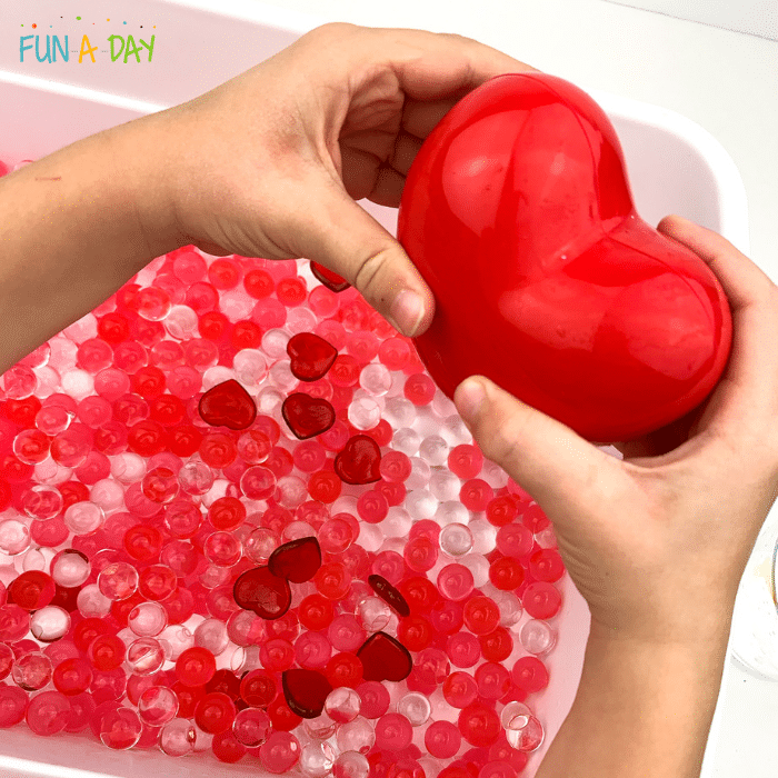 child's hands holding heart container above valentine water beads bin