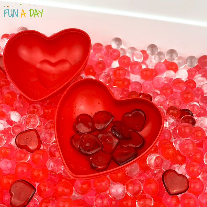 Red acrylic hearts within a heart container inside a valentine sensory bin of clear and red water beads