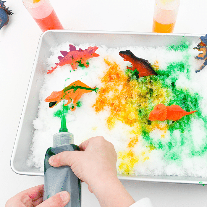 Preschool hands squeezing green snow paint on dinosaurs in snow tray.