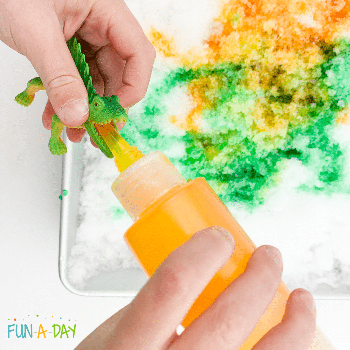 Preschool hands holding dinosaur toy and bottle of yellow snow paint over snowy sensory bin.