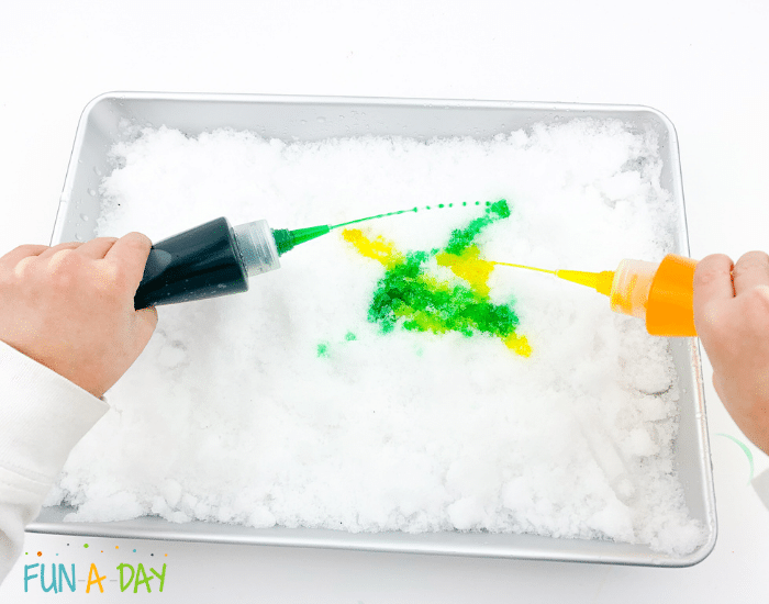 Preschooler using both hands to squeeze green and yellow snow paint on tray of snow.