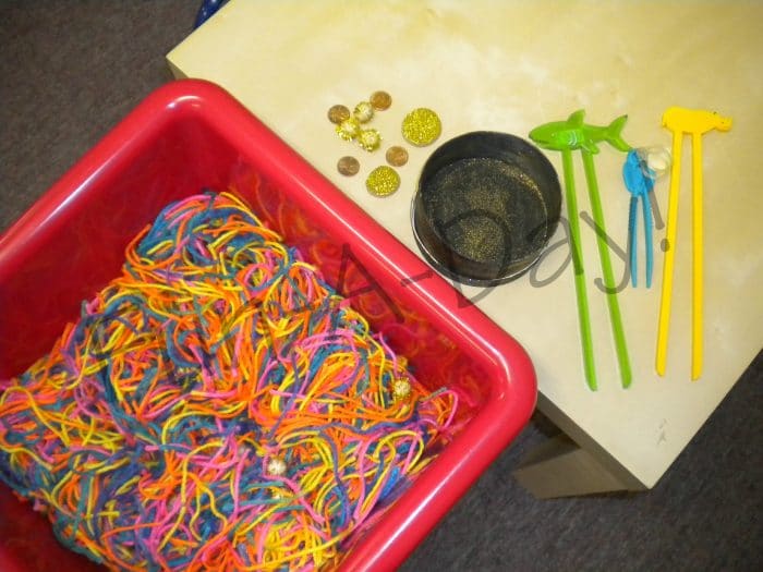 rainbow yarn in a sensory bin, with chopsticks, gold coin manipulatives, and small black box nearby