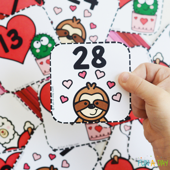 pile of number cards for valentine calendar with child's hand holding the number 28 with a cute sloth face on it