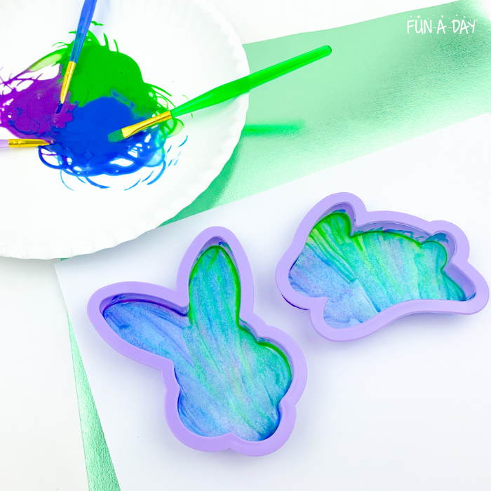 Paint brushes sitting on plate of mixed purple, green, and blue paints next to a paper where cookie cutters have been used as stencils to paint bunny shapes.