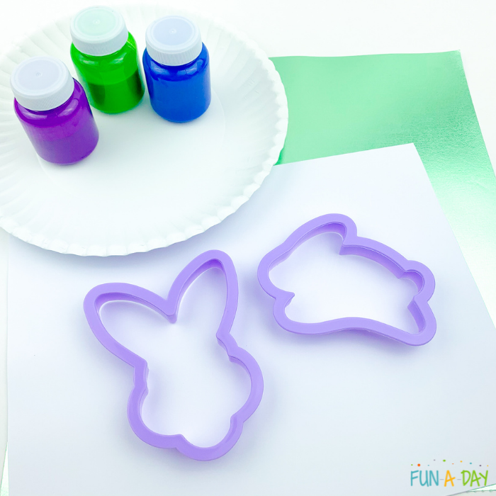Materials from the Easter cookie cutter art which include small bottles of purple, green, and blue paints, a paper plate, paper, and bunny-shaped cookie cutters.