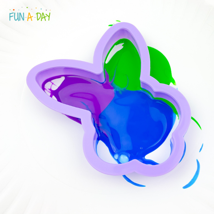 Bunny cookie cutter being dipped into purple, green, and blue swirled paint.