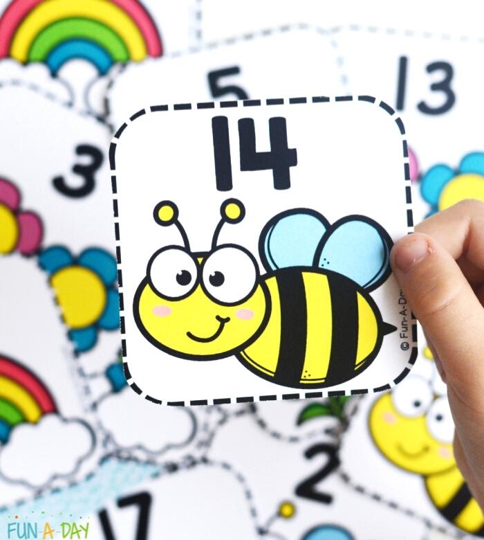 Child's hand holding bee card with number 14 in foreground and a pile of spring number cards in the background.