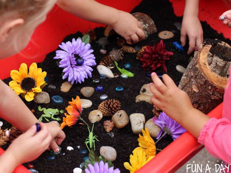 Kids playing in garden messy play sensory bin filled with dirt, rocks, pine cones, toys, and glass gems.