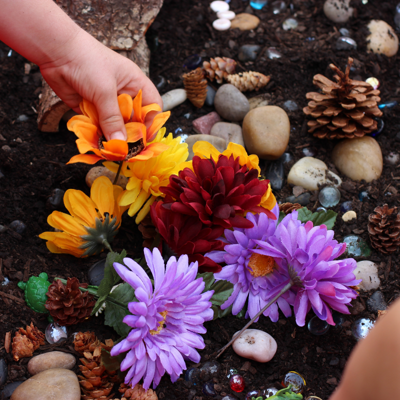 Preschooler's hand placing fake flowers in a garden messy play sensory bin filled with dirt and rocks.