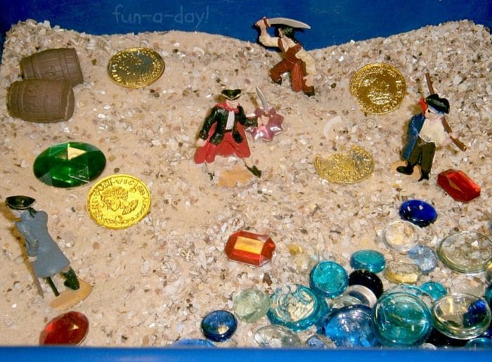 Close up of sandy sensory bin with pirate toys, gold coins, and glass gems