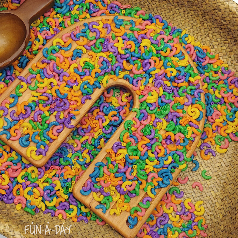 Colorful dyed elbow macaroni in a bin with wooden rainbow tray.