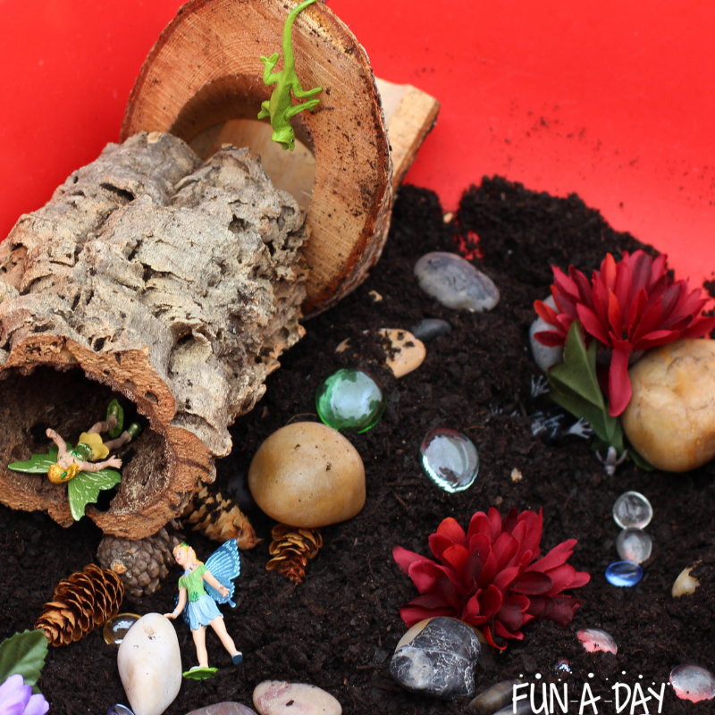Wood pieces, fairy toys, chameleon toy, fake flowers, rocks, and glass gems in a sensory bin of dirt.