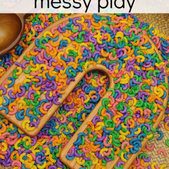 Rainbow dyed macaroni covering rainbow-shaped wooden tray. With text that reads rainbow messy play.