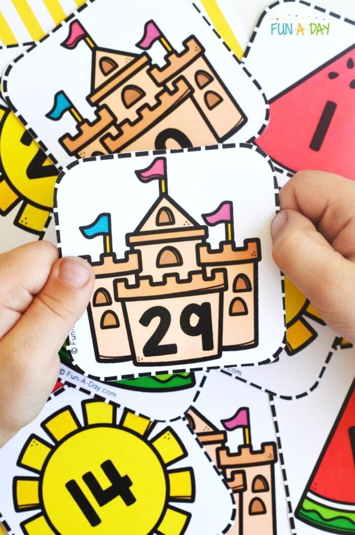 child's hands holding a sand castle card with the number 29 on it, with more number cards in the background
