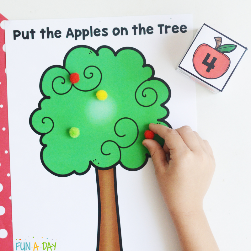 Preschooler's hand adding pompoms to apple tree math game printable