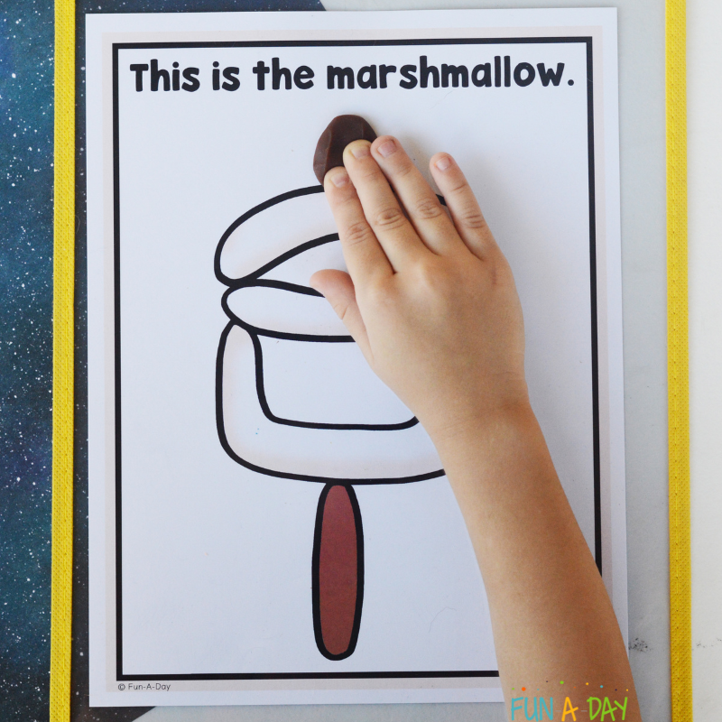 preschooler's hand placing brown playdough on camping playdough mat that reads this is a marshmallow
