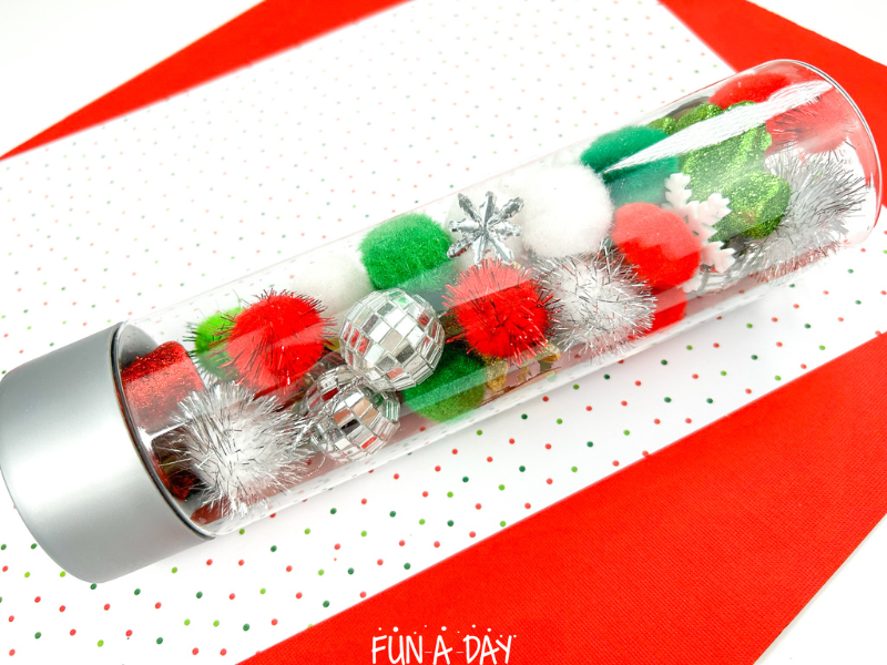 christmas sensory jar on its side, filled with pom poms, snowflakes, and small ornaments