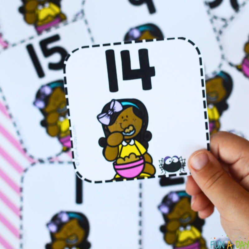 Holding the Little Miss Muffet number card number 14 above the other cards splayed out on a table.